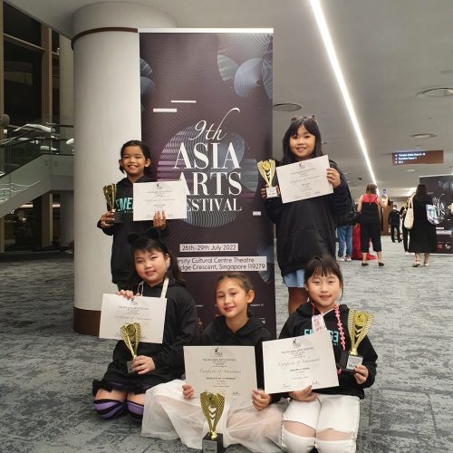 Five young girls in black hoodies kneeling and standing proudly with trophies and certificates in front of an Asia Arts Festival banner