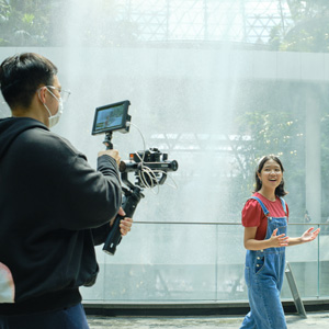 Videographer holding a gimbal camera rig filming a young girl speaking and gesturing in front of an indoor waterfall