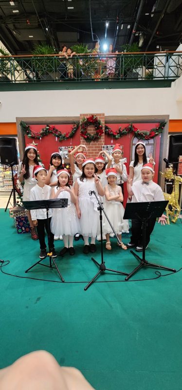 A group of children dressed in festive white outfits and Santa hats pose on stage in front of a Christmas-themed backdrop