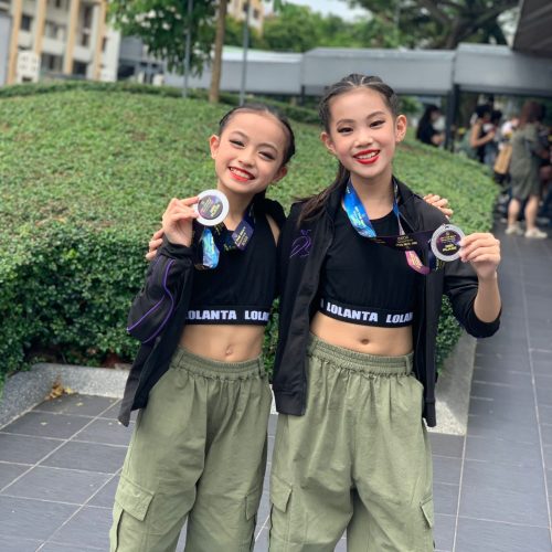 Two young girls in matching Lolanta tops and green cargo pants smiling proudly while holding their medals outdoors