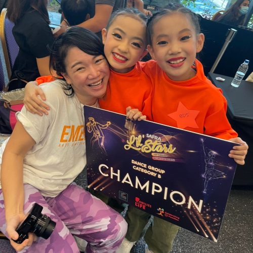 Two smiling young dancers in orange costumes and their dance coach hugging tightly while holding their Category B champion board