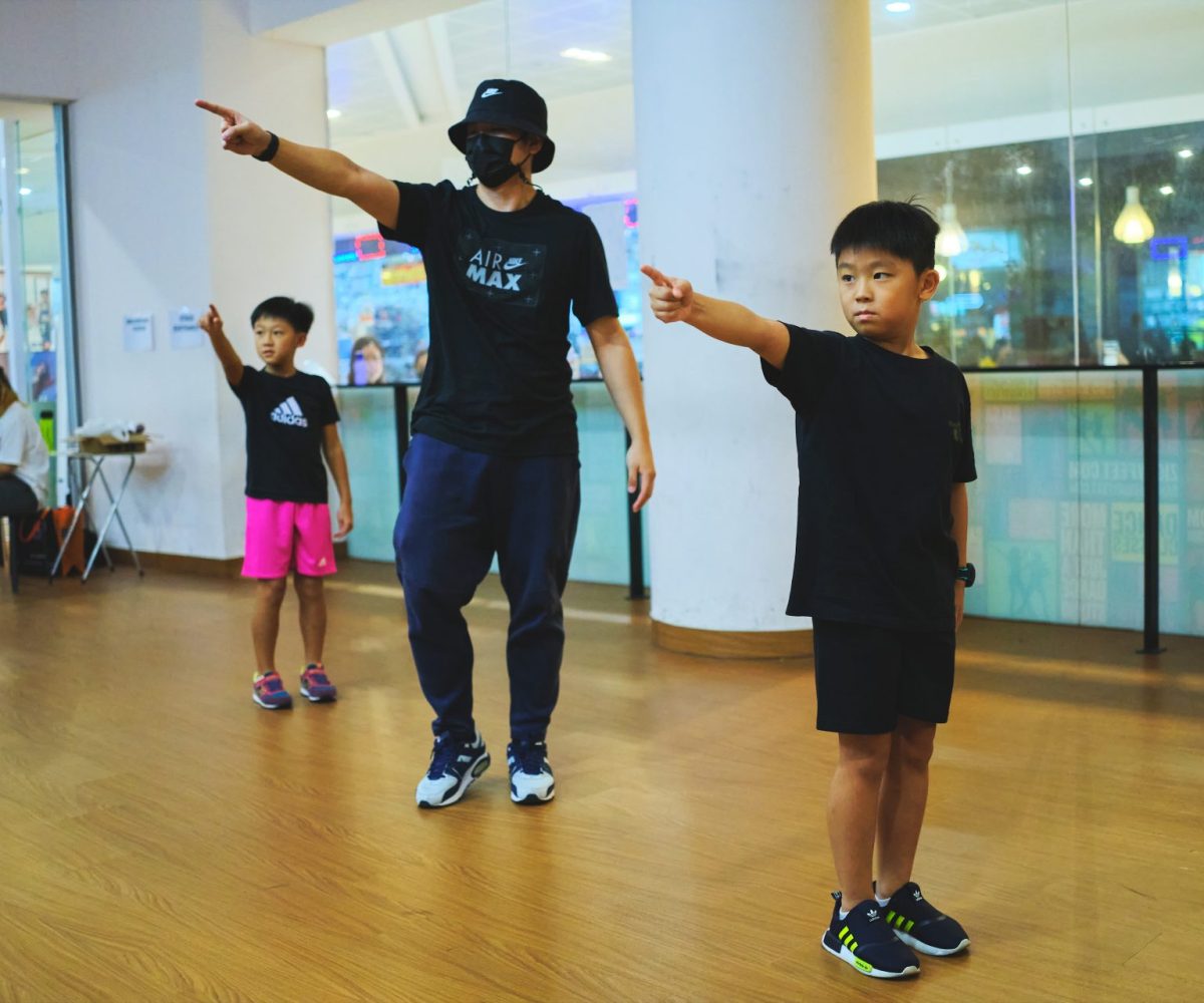 Dance instructor in a bucket hat and mask guiding young boys through pointing dance movements in a bright studio