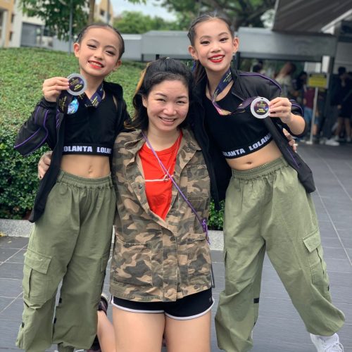 Two smiling young dancers in green cargo pants holding up their competition medals beside their proud coach outdoors