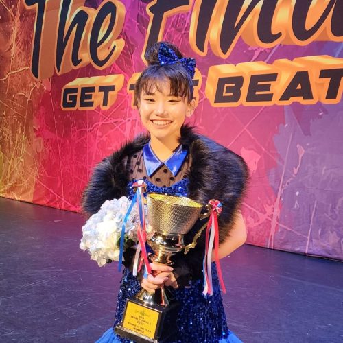 Young dancer in a royal blue sequin dress holding a champion cup and bouquet of flowers against a vibrant backdrop