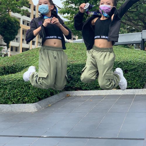 Two young dancers wearing face masks and olive cargo pants jumping high into the air while holding their medals