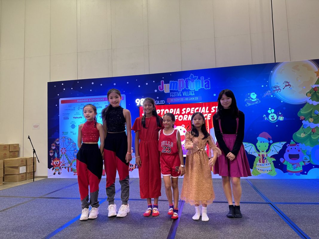 Six young girls in colorful outfits pose together on a stage in front of a Jumptopia event backdrop