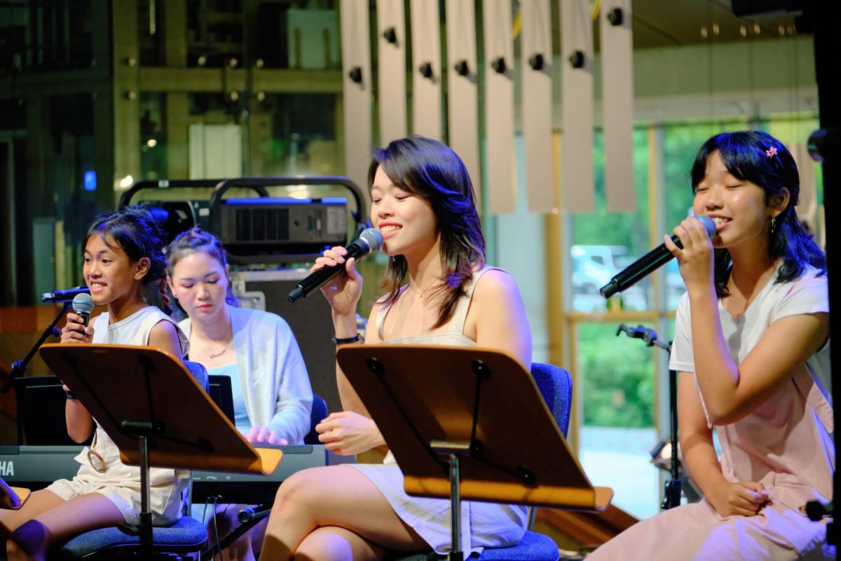 Three young women sit on stage holding microphones and singing, accompanied by a pianist in the background