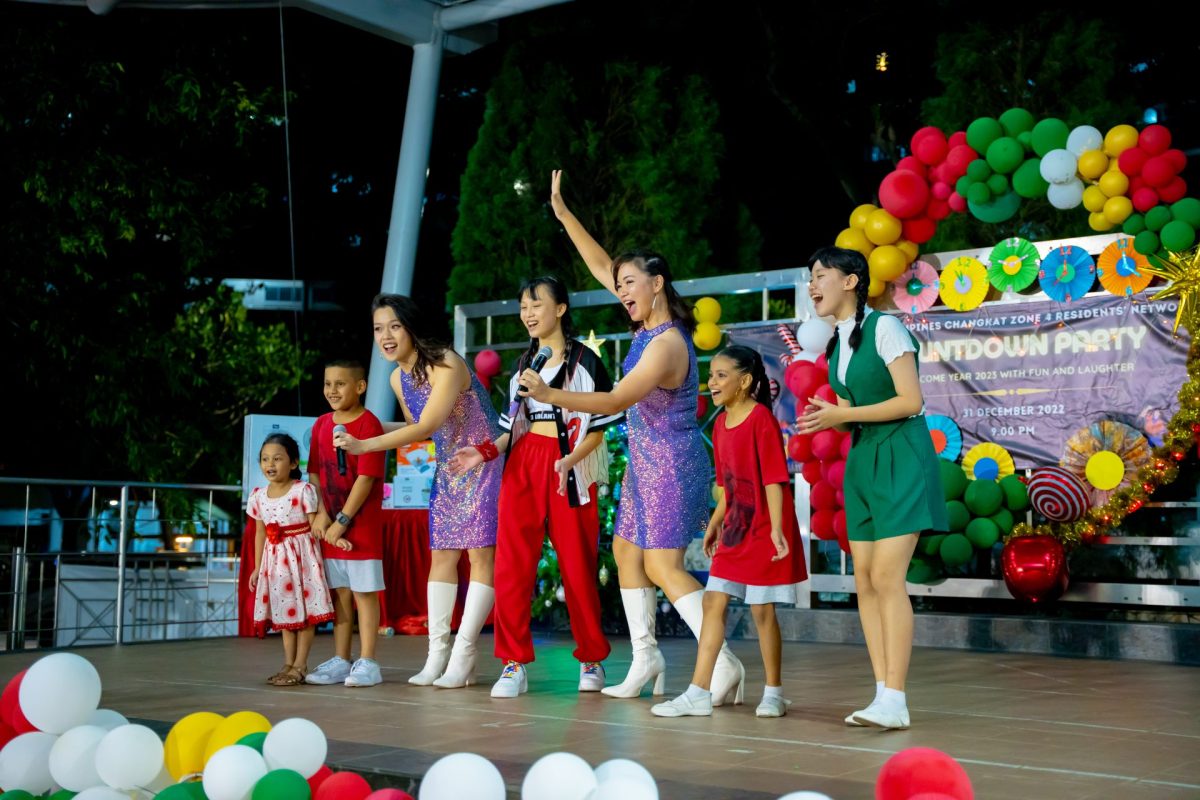 Performers in festive outfits sing and dance on an outdoor stage decorated with colorful balloon arches during a countdown party