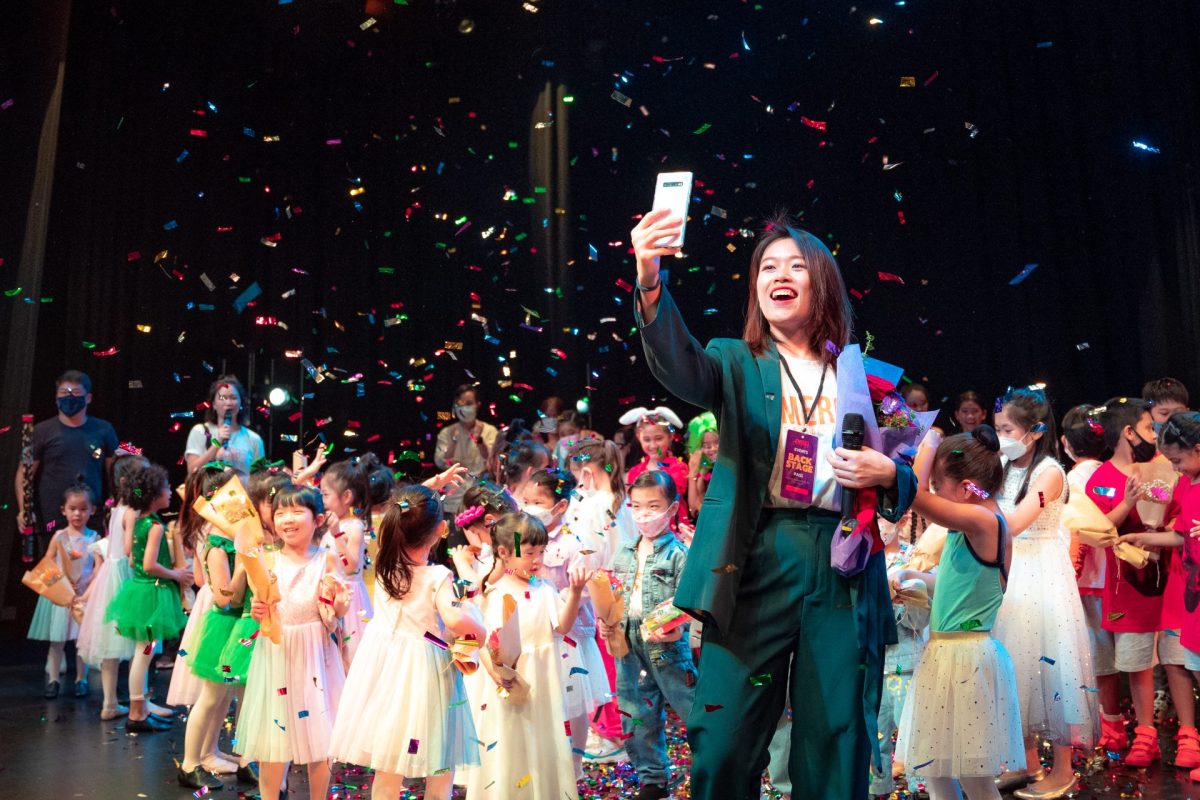 A woman in a green suit takes a selfie on stage with young performers as colorful confetti falls around them