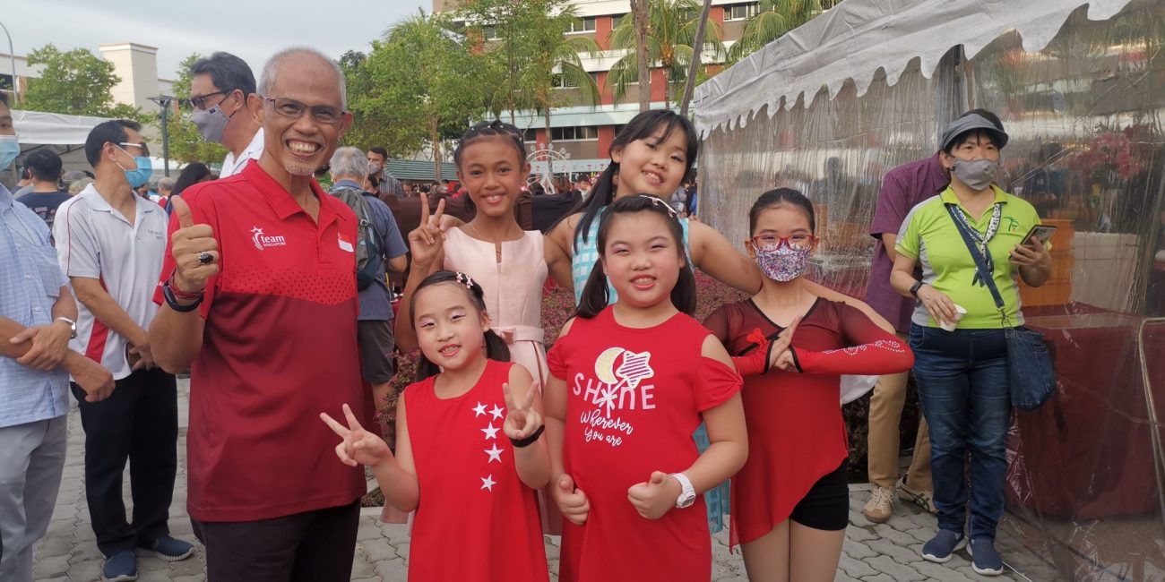 A smiling man giving a thumbs-up poses outdoors with a group of young girls and community members dressed in red and white