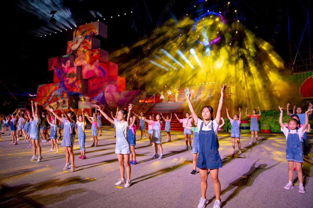 A large group of young performers wearing denim overalls reach their hands toward the sky during a vibrant outdoor stage production lit by colorful spotlights