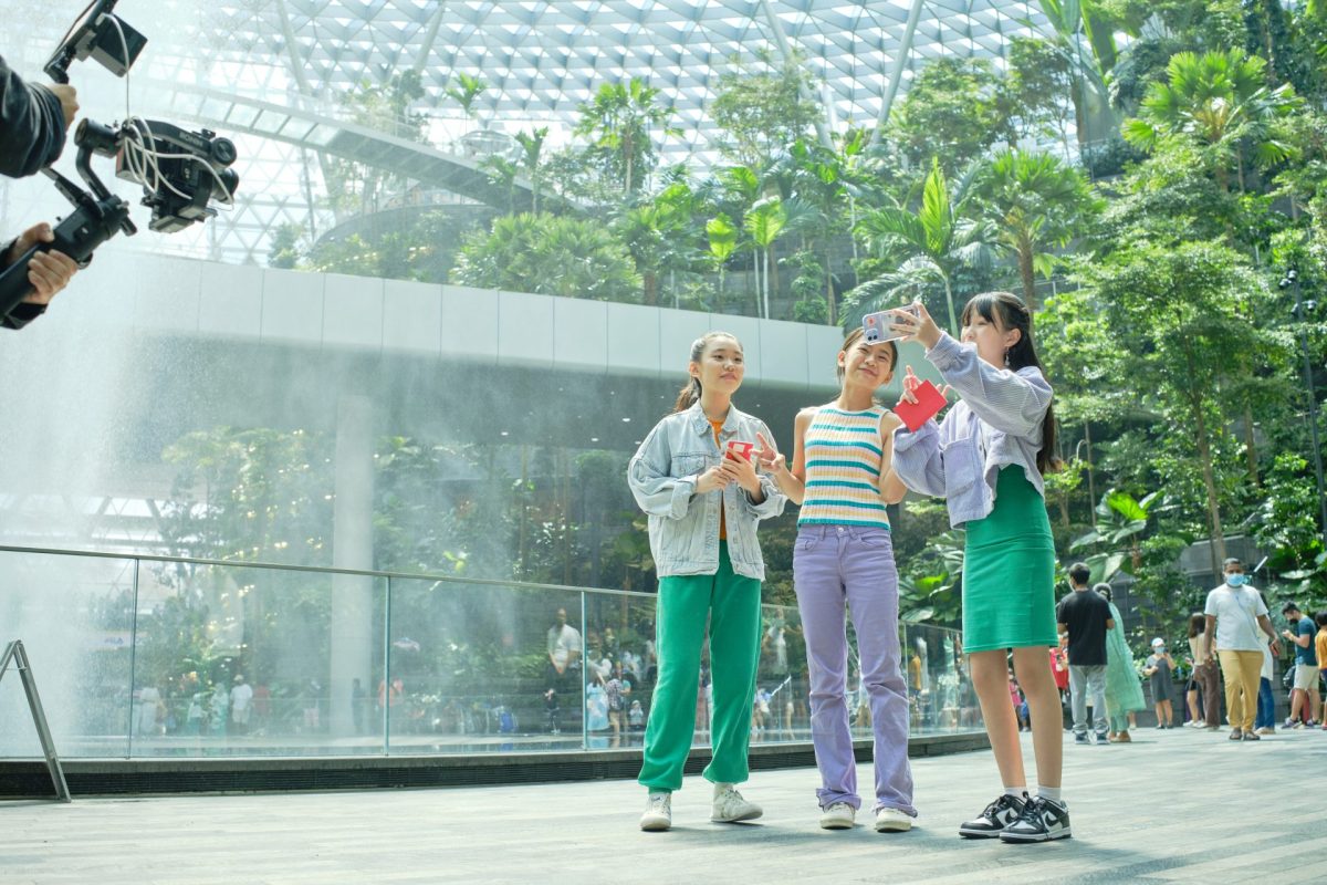 Three young girls take a selfie inside a lush indoor greenhouse setting near a waterfall