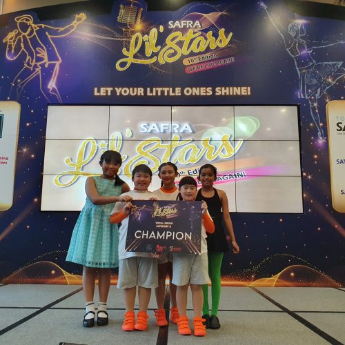 Five young performers in costumes standing side by side holding their Vocal Group Category B Champion board on a lit event stage