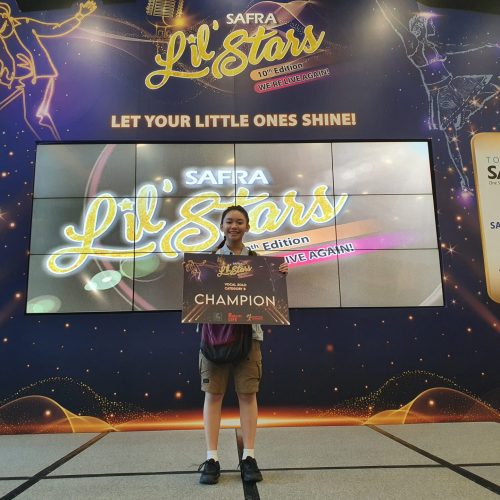 Young girl standing on stage proudly presenting her Vocal Solo Category B Champion award board at the SAFRA Lil Stars competition