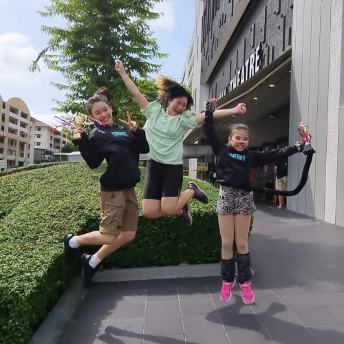 Three young performing arts students leaping simultaneously into the air while holding their winning trophies outside a venue