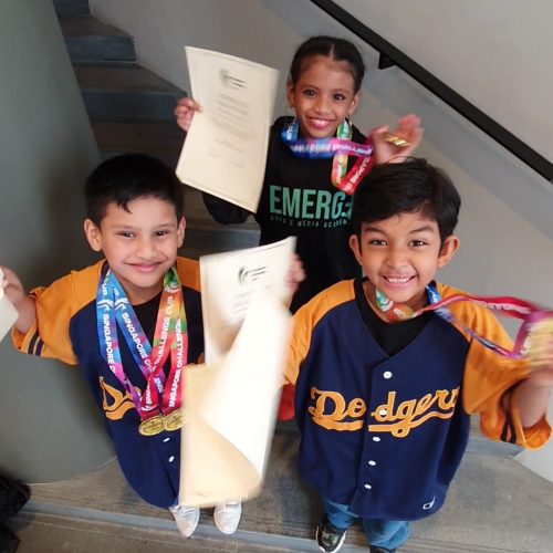 High-angle shot of three cheerful young performers smiling up toward the camera while waving their award certificates and medals