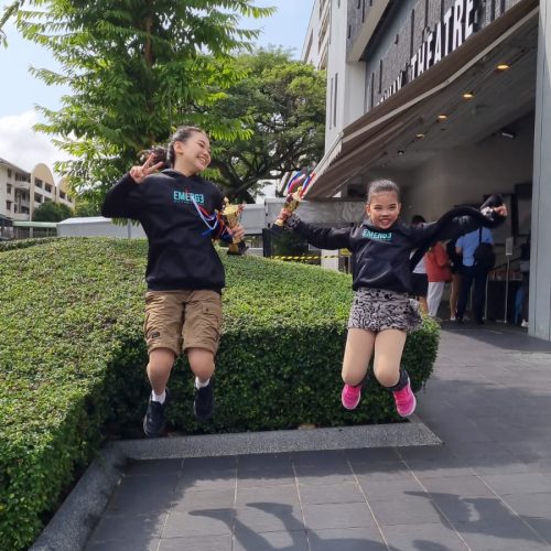 Two excited girls wearing black Emerge hoodies leaping into the air while holding competition trophies outside a theatre