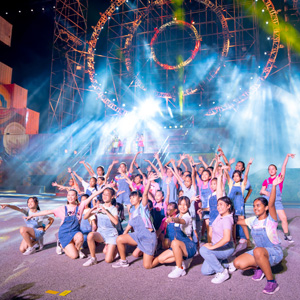 Large ensemble of smiling children in denim overalls posing with arms raised under bright concert lights and theatrical smoke