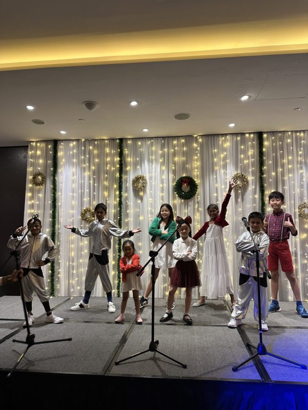 A group of children strike energetic poses on a stage in front of a fairy-light backdrop decorated with Christmas wreaths