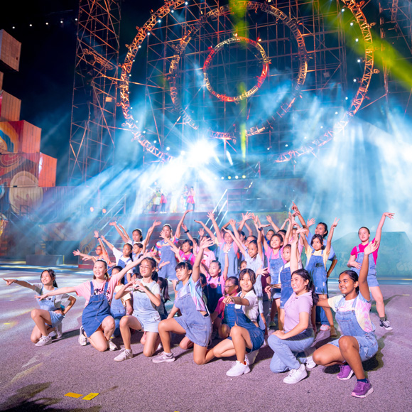 Large ensemble of smiling young dancers in denim outfits raising their arms together on an illuminated concert stage with smoke effects