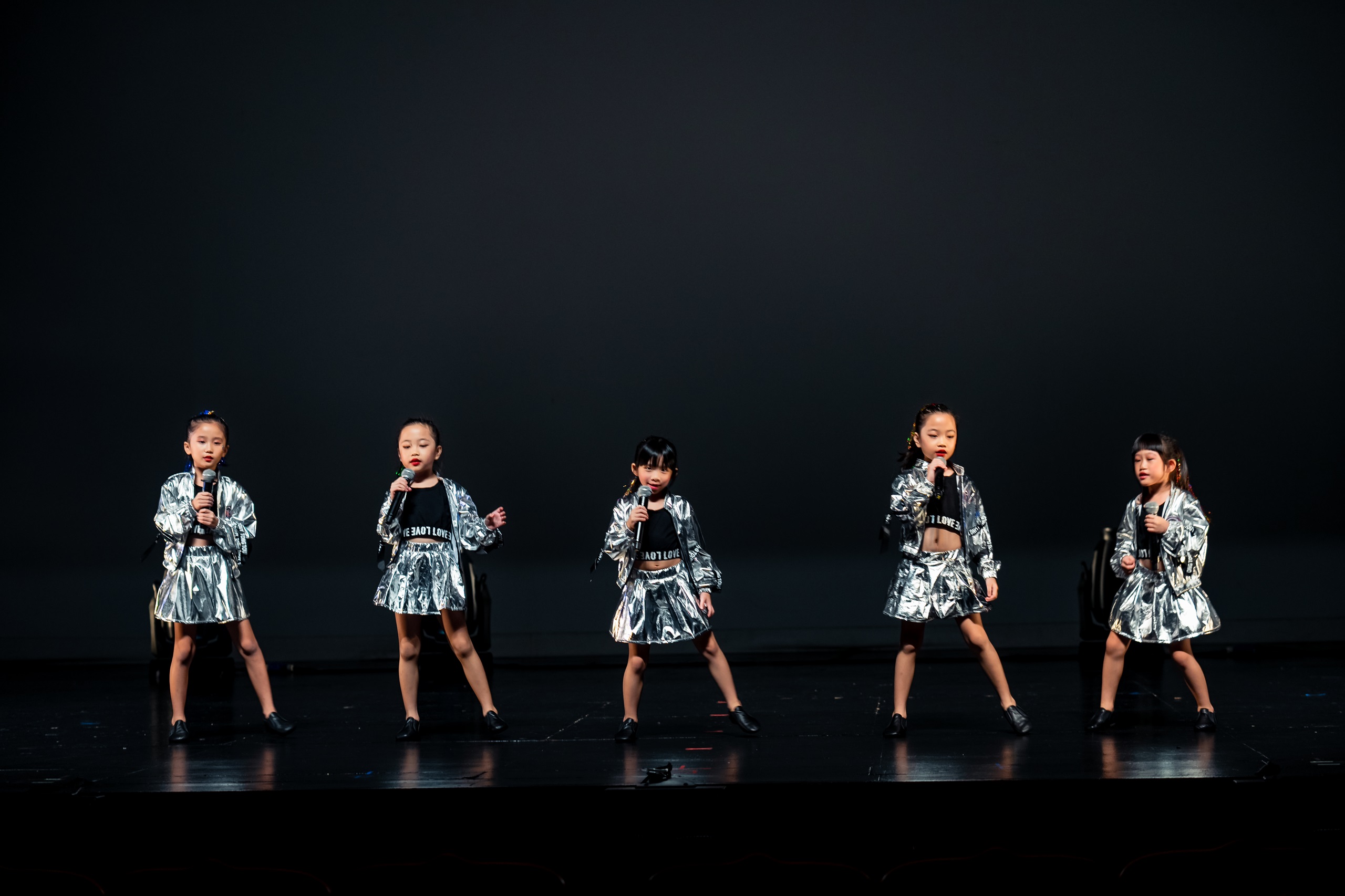 Five young girls in matching metallic silver jackets and skirts hold microphones while performing on a dark stage