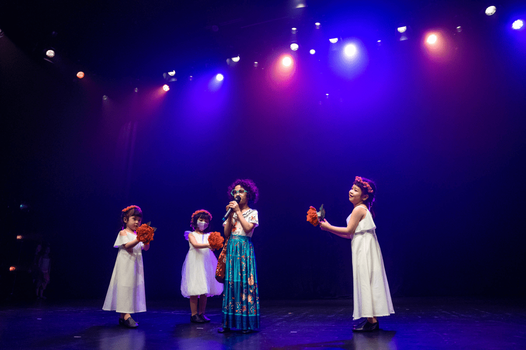Four young performers on a theater stage under purple spotlights including a lead singer in floral skirt surrounded by girls in white dresses holding flowers