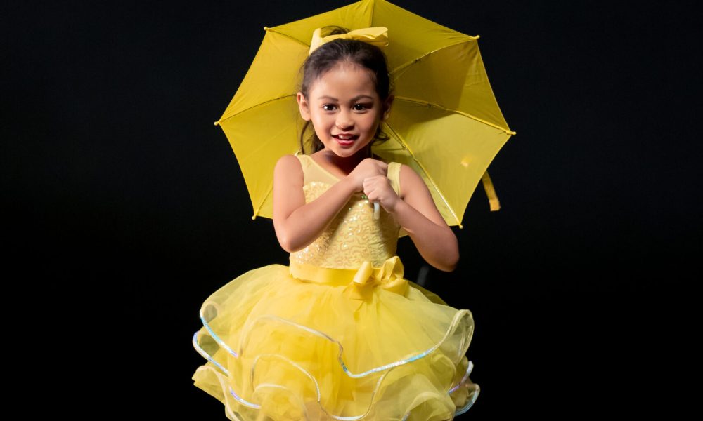 Smiling young girl in a yellow tiered tutu holding an open yellow parasol on a dark theater stage