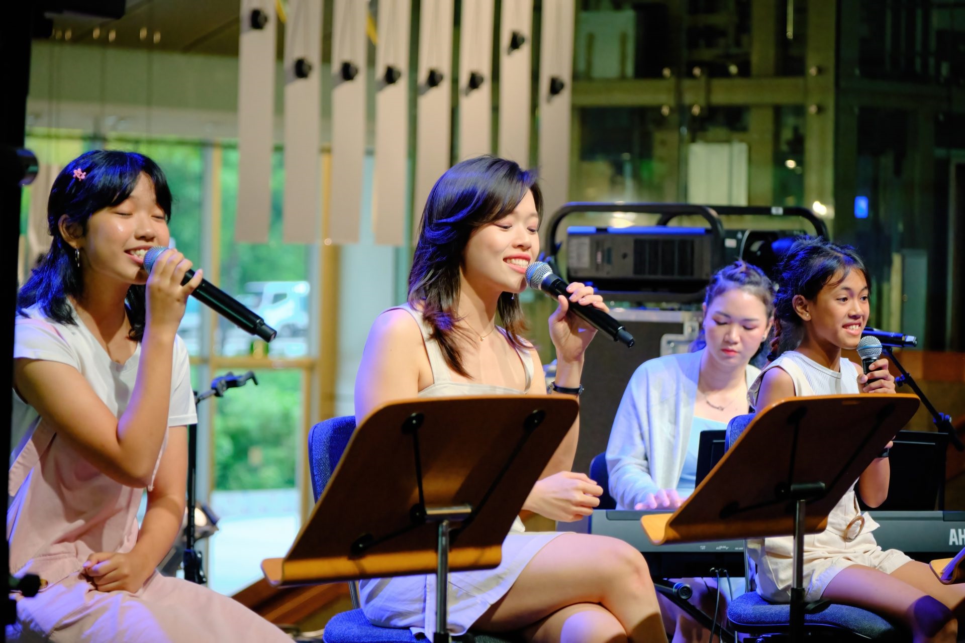Three singers perform into microphones on stage while seated behind music stands, accompanied by a female pianist in the background