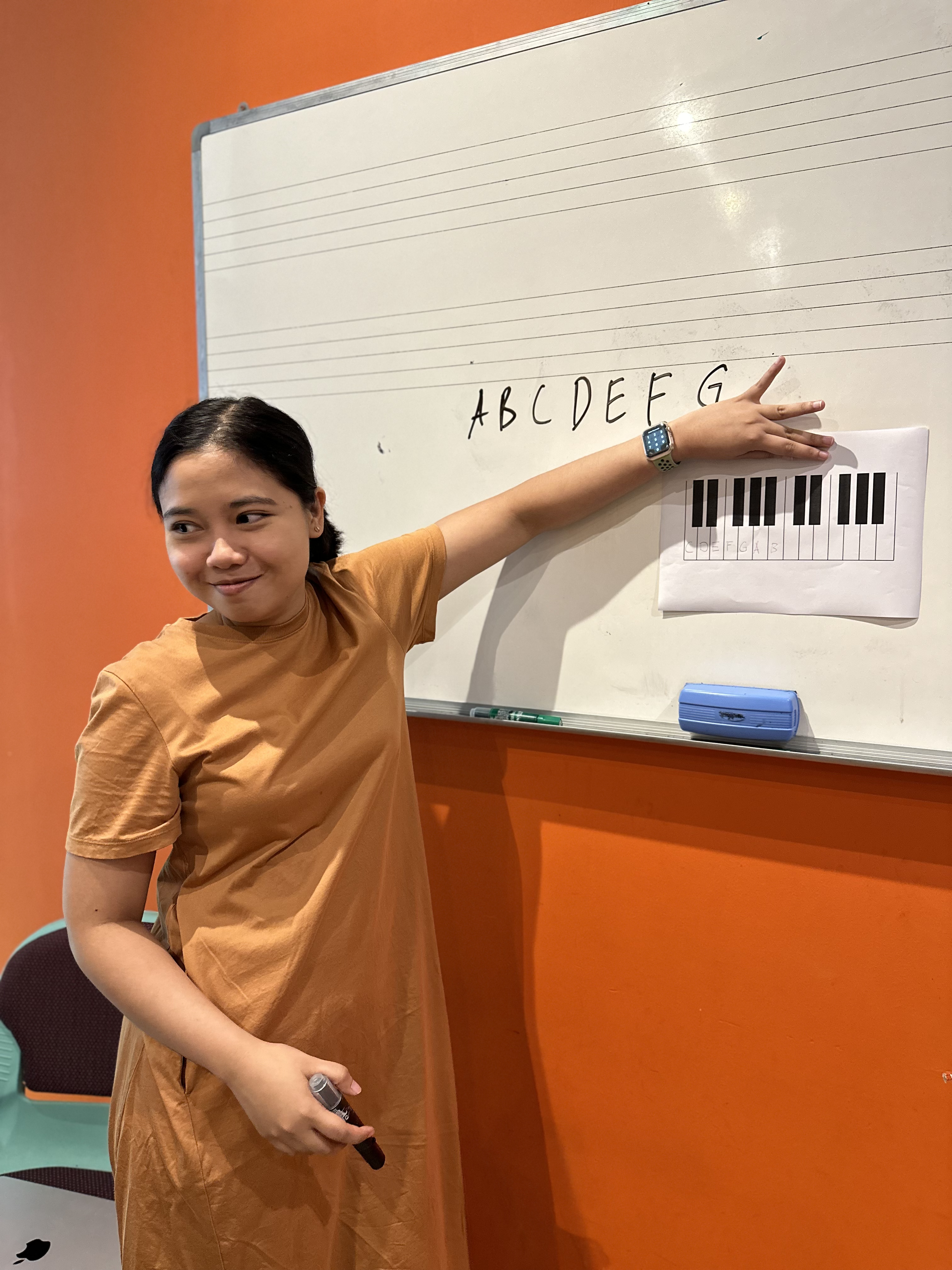 A music teacher in a brown dress points to musical notes and a keyboard diagram on a whiteboard