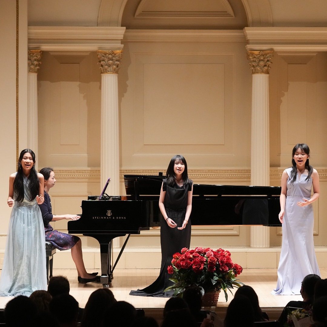 Three female singers in elegant formal gowns perform on stage beside a black Steinway grand piano and a pianist