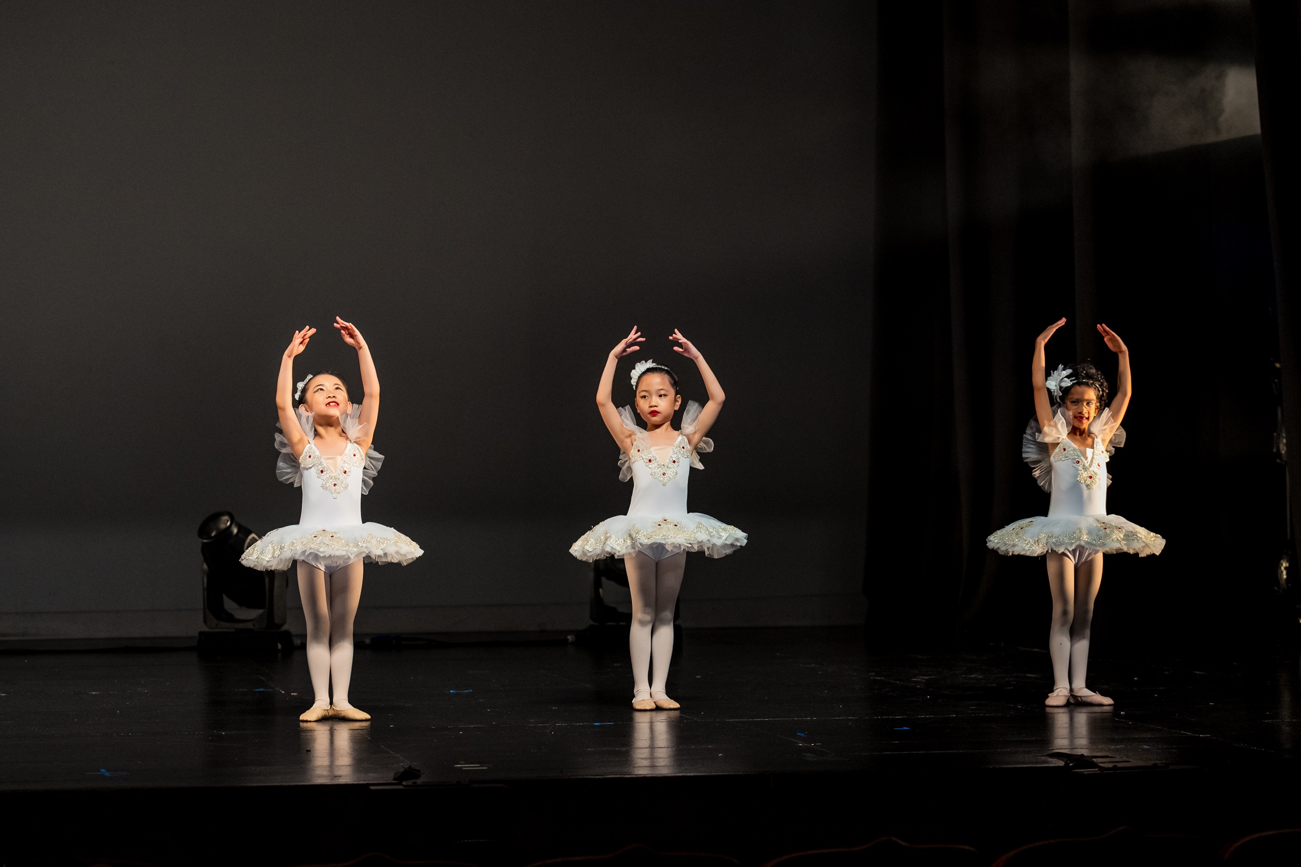 Three young girls in matching white tutus perform a ballet pose on stage