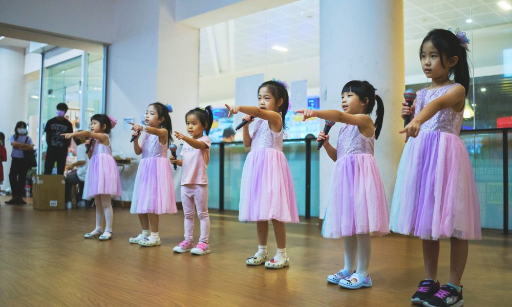 Line of young girls wearing lavender tulle dresses singing into handheld microphones with choreography gestures in a dance studio