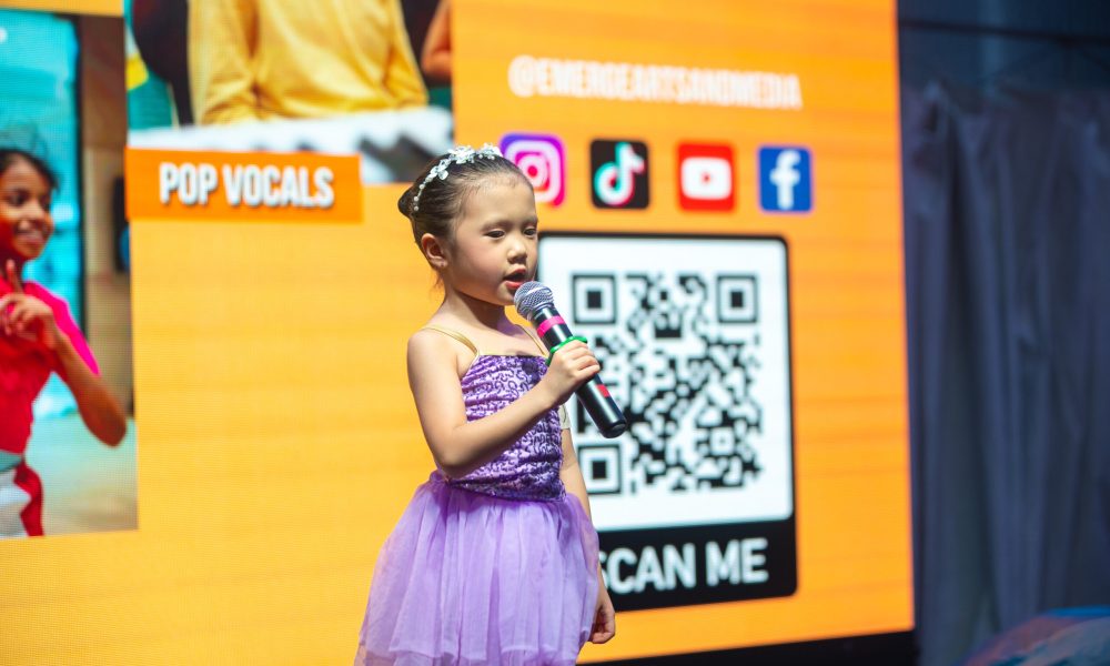 Young girl in a purple dress singing into a microphone in front of a yellow digital screen with QR code and pop vocals text