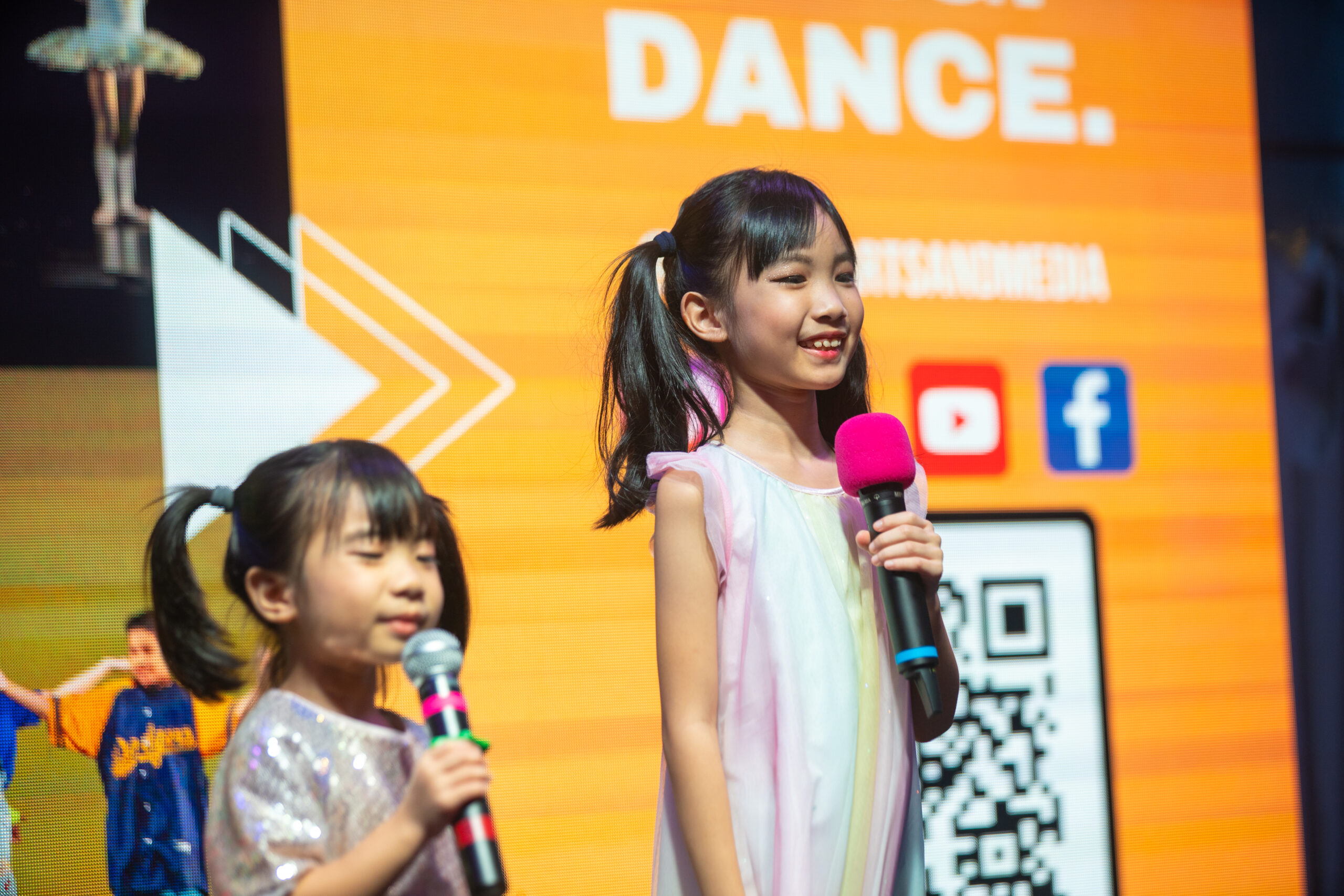 Two young girls with twintails holding microphones and speaking on stage in front of a bright orange banner