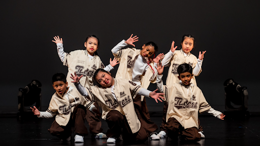 Six children wearing matching cream-colored Tigers jerseys and brown pants pose playfully on stage with open hands during a hip-hop dance routine