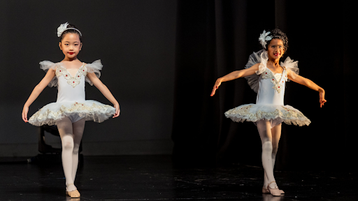 Two young girls in ornate white ballet tutus and headpieces performing a ballet routine on a dark stage