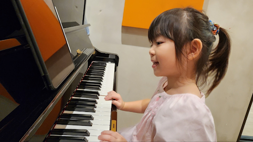 A young girl with a ponytail in a light pink dress smiles while playing a black upright piano