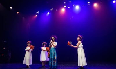Young girl singing into a microphone in an Encanto style costume surrounded by three girls in white dresses holding flower bouquets under colorful stage lighting