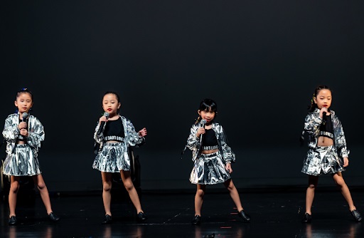 Four young girls wearing shiny metallic silver jackets perform a song while holding microphones on stage