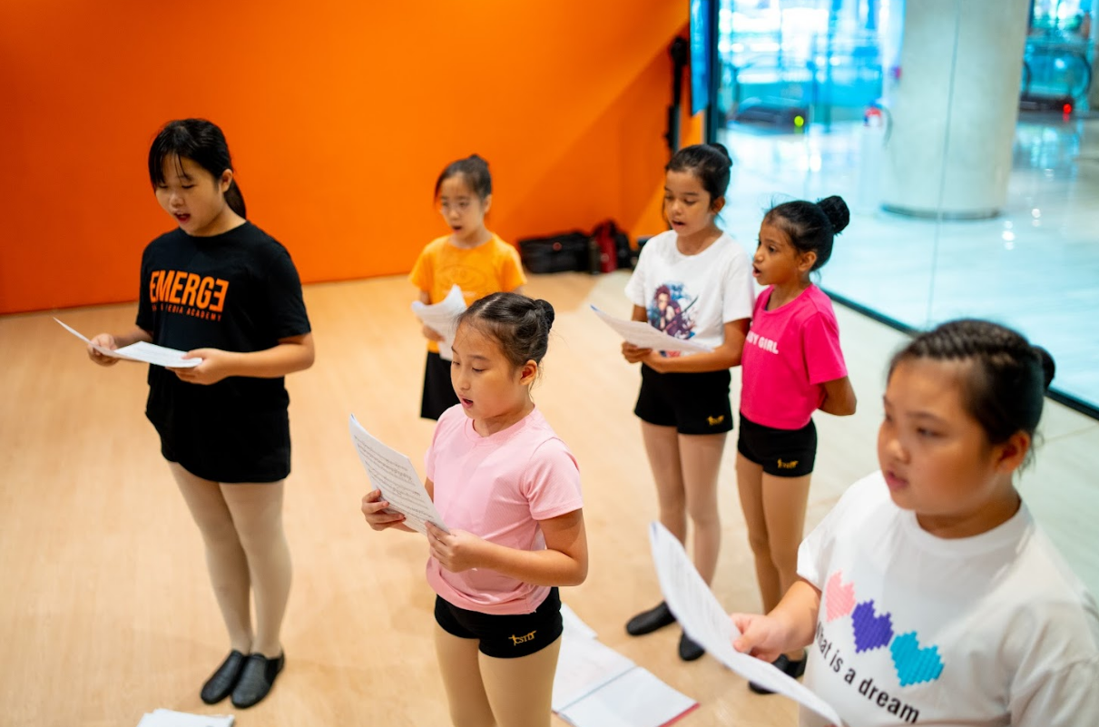 A group of young girls holding papers and reading lyrics while practicing singing together in a brightly lit studio room