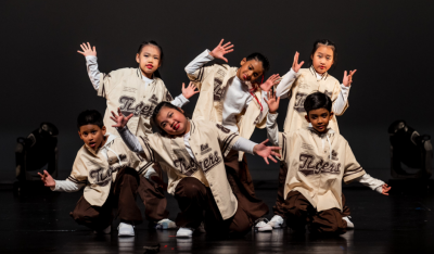 kids-hip-hop-dance-crew-matching-baseball-jerseys-stage-pose Group of diverse children posing together in matching oversized baseball jerseys and brown pants during a hip-hop dance performance on stage