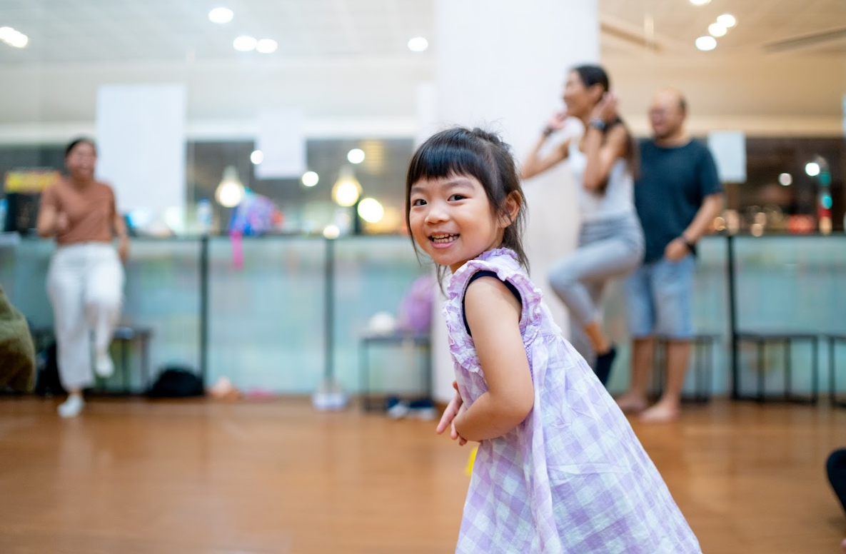A young girl in a purple patterned dress smiles at the camera while moving around a spacious wooden-floored dance studio with adults in the background