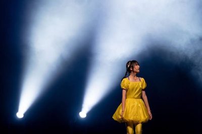 Young girl in a bright yellow puff dress standing in theatrical haze under two angled spotlights on a dark stage