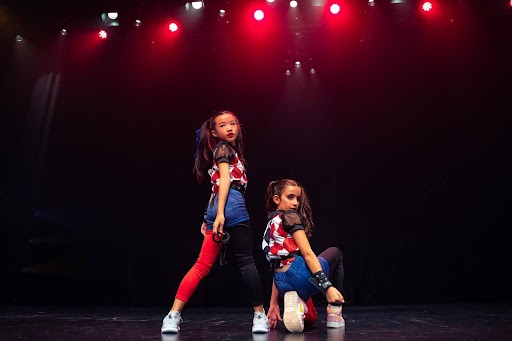 Two young girls in matching red, blue, and black outfits strike stylish, dynamic poses under red stage spotlights