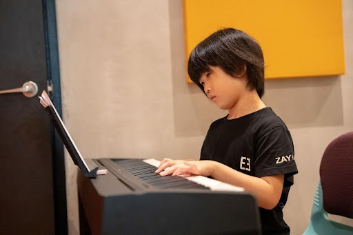 A young boy wearing a black shirt looks closely at sheet music while playing a digital piano in a lesson studio
