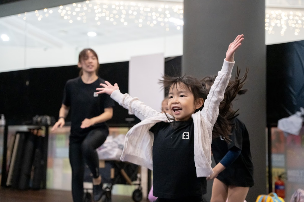 A joyful young girl wearing a white cardigan throws her arms up wide while dancing in a studio class with an instructor in the background