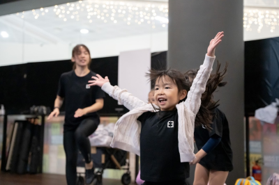 young-girl-expressively-dancing-in-studio-class A joyful young girl wearing a white cardigan throws her arms up wide while dancing in a studio class with an instructor in the background