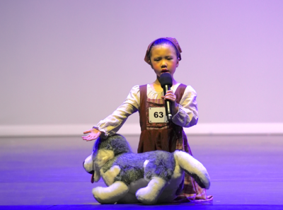 young-girl-performing-onstage-with-plush-dog-prop A young girl in a vintage-style brown dress and headscarf sings into a microphone while petting a stuffed husky dog prop on stage