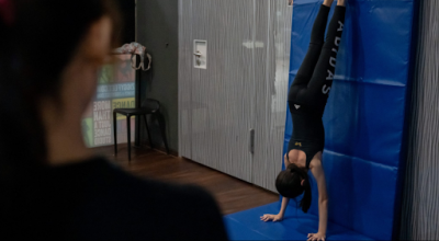 young-girl-practicing-handstand-against-wall-mat A young girl in black activewear performs a handstand against a blue safety mat under the supervision of an instructor in a dance studio