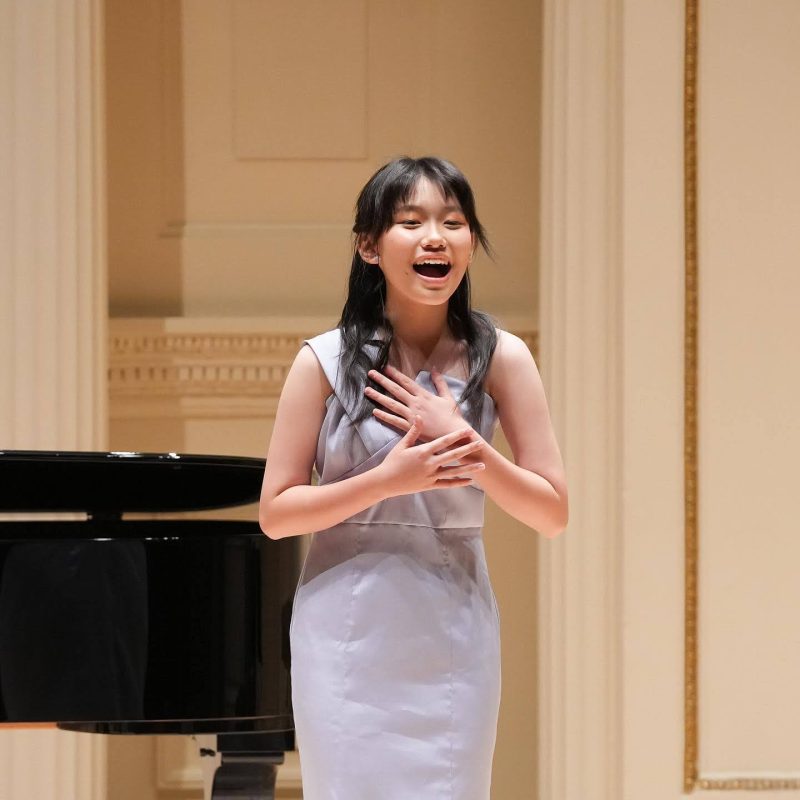 A young girl in a light blue dress sings passionately with her hands resting on her chest next to a grand piano on stage